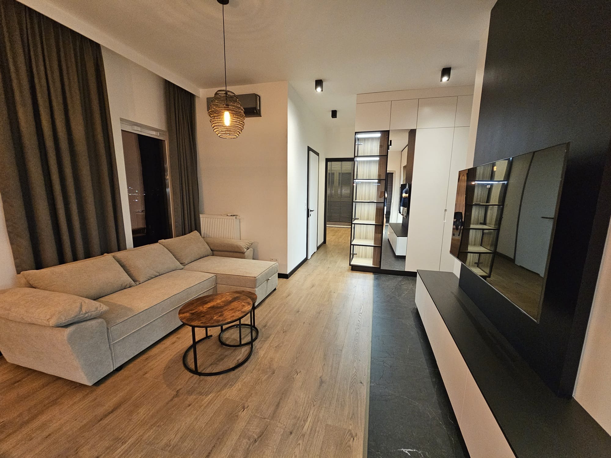 Nowoczesny salon z beżową sofą narożną, okrągłym stolikiem kawowym z drewnianym blatem, podłogą z paneli imitujących drewno i dekoracyjną lampą wiszącą z plecionki.