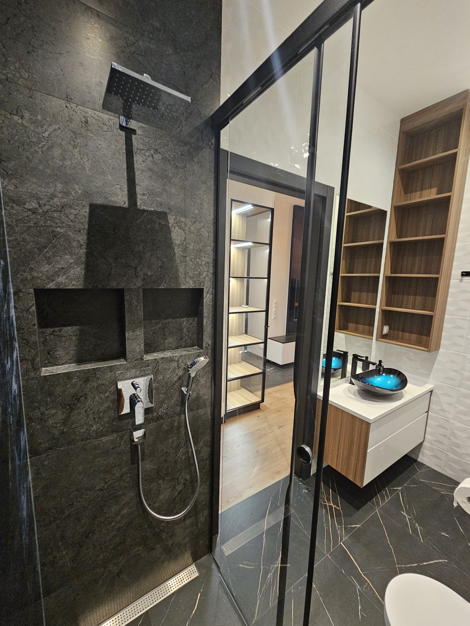 Nowoczesna łazienka z ciemnymi płytkami imitującymi kamień na ścianie prysznicowej, wbudowanymi półkami, deszczownicą i odpływem liniowym. W tle widoczna toaletka z błękitną umywalką oraz regał...
