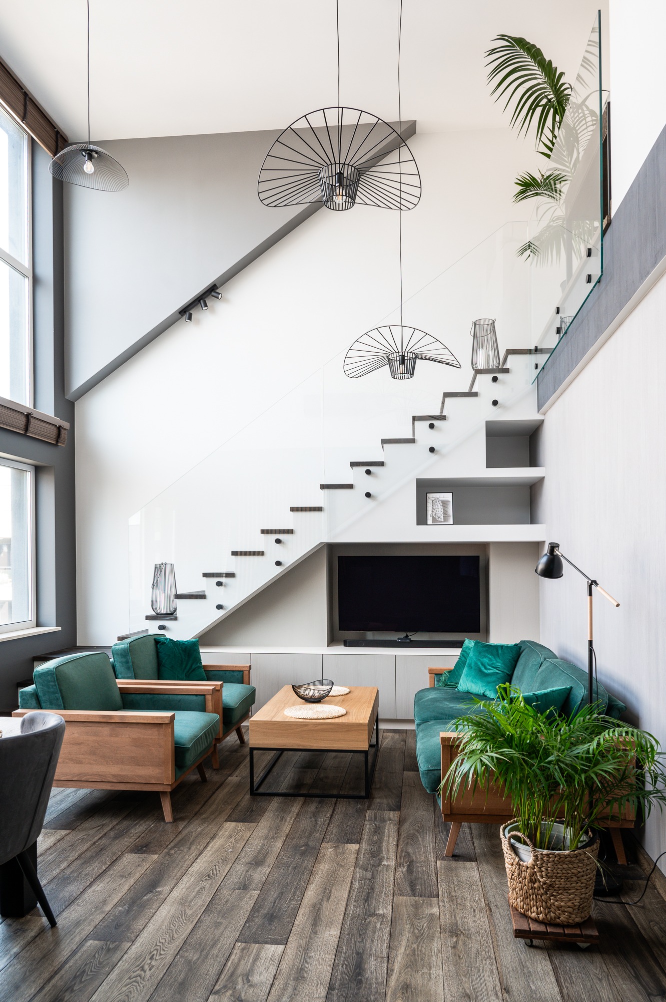 Jasne, nowoczesne wnętrze salonu z drewnianą podłogą, zielonymi sofami i fotelami, schodami z szklaną balustradą i designerskimi lampami wiszącymi.