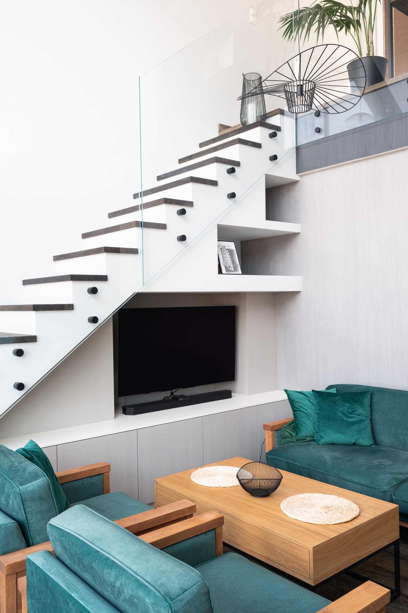 Nowoczesne schody z białymi stopnicami i szklaną balustradą, wkomponowane w aranżację salonu z turkusowymi meblami i drewnianym stolikiem kawowym. Pod schodami zabudowa z telewizorem i soundbarem.