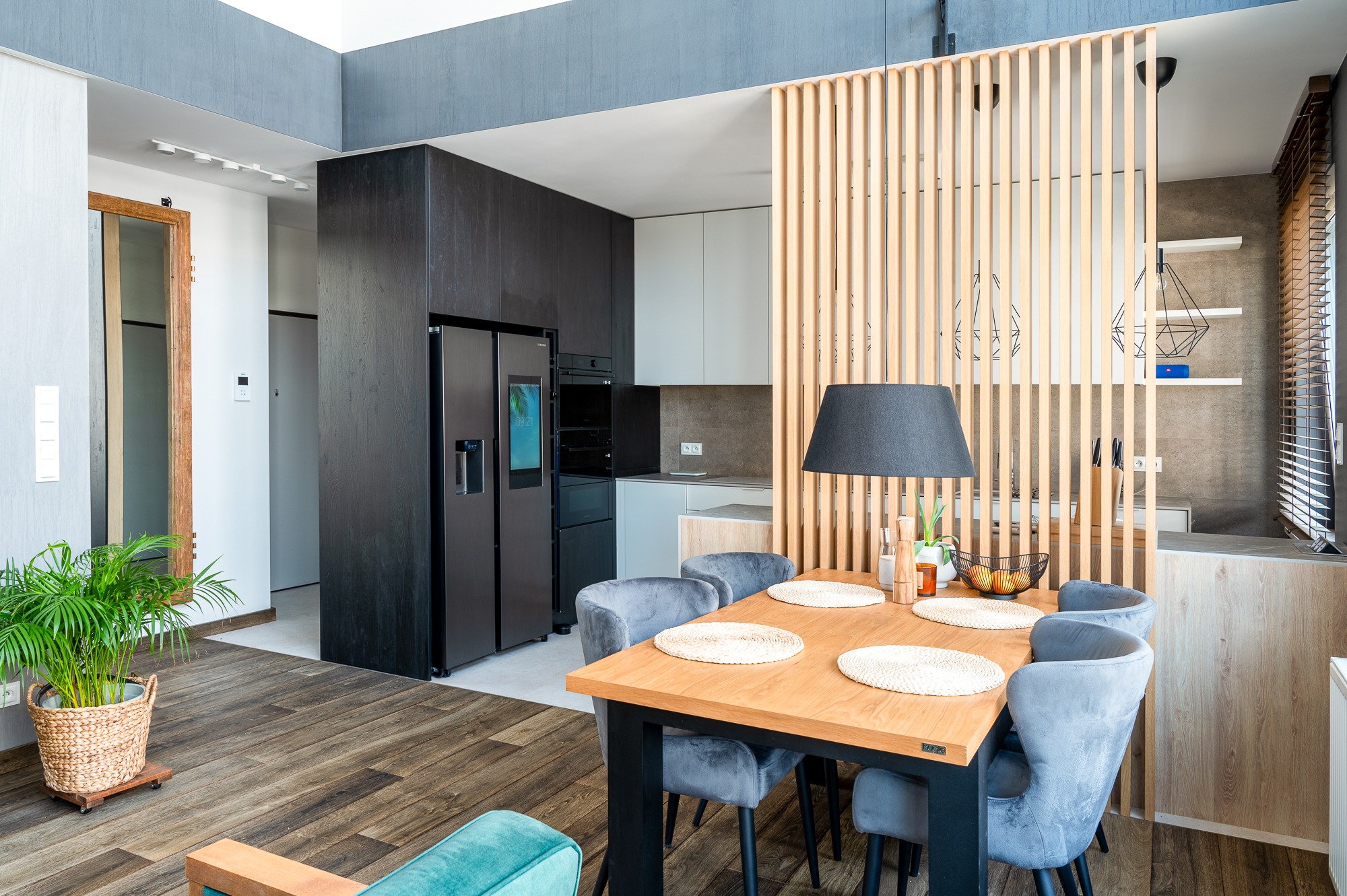 Nowoczesna jadalnia z drewnianym stołem, szarymi krzesłami i ażurową ścianką działową z jasnego drewna, widoczna kuchnia w tle.