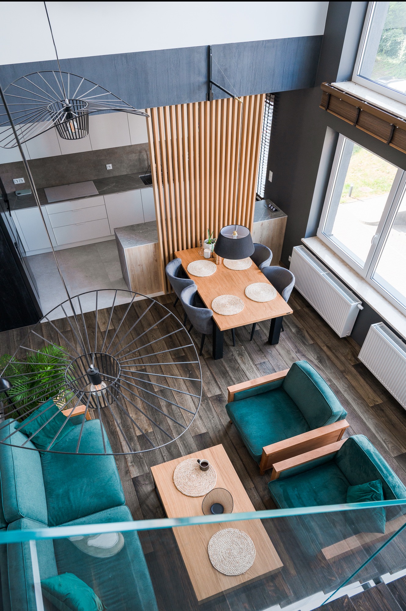 Nowoczesne wnętrze z perspektywy góry: salon z turkusową sofą i drewnianymi stolikami kawowymi, jadalnia z drewnianym stołem i szarymi krzesłami, kuchnia w bieli i szarości, oddzielona drewnianą...