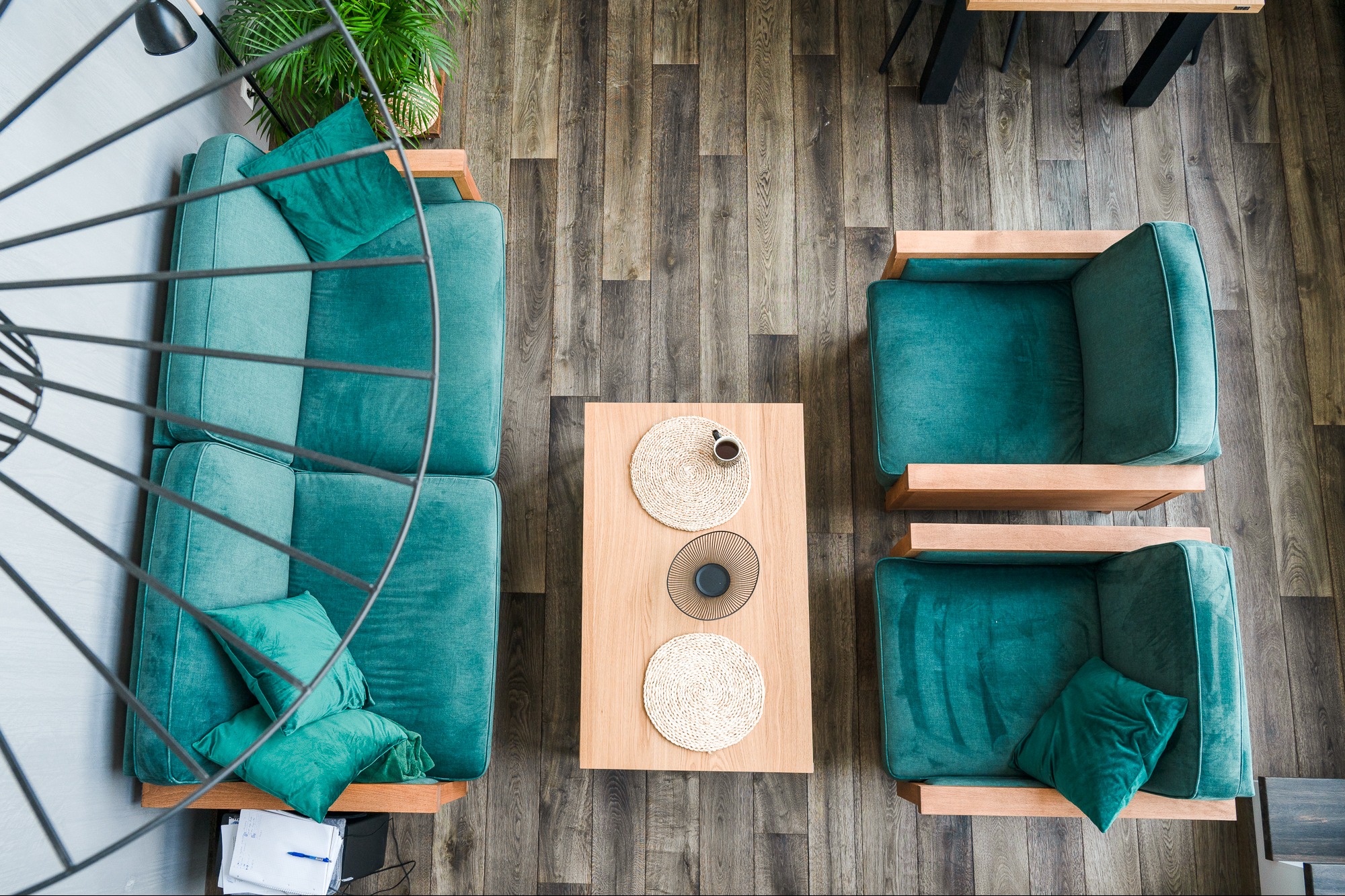 Widok z góry na salon z drewnianą podłogą, turkusową sofą i fotelami z drewnianymi ramami, stolik kawowy z dekoracjami i filiżanką, oraz fragment metalowej lampy.