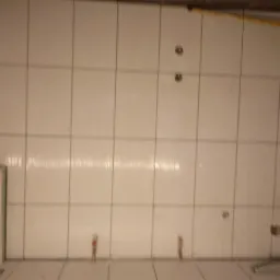 Ściana łazienki wyłożona białymi płytkami, widoczne otwory na armaturę, fragment płyty gipsowej przy ścianie, żółty kabel pod sufitem.