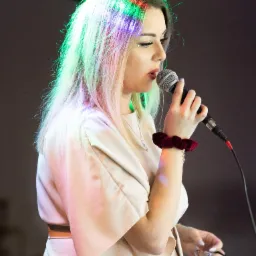 Wokalistka z blond włosami, oświetlonymi kolorowymi światłami, śpiewa do mikrofonu, ubrana w beżową kreację z paskiem, z bordową gumką na nadgarstku i delikatnym makijażem.