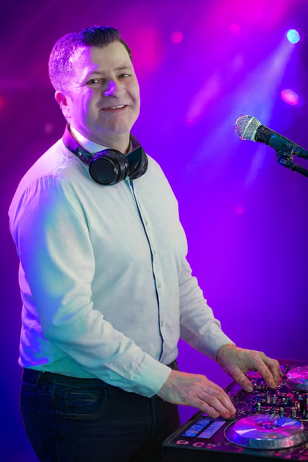 Uśmiechnięty DJ w białej koszuli, z słuchawkami na szyi, miksuje muzykę na konsolecie pod fioletowym oświetleniem scenicznym. Mikrofon po prawej stronie.