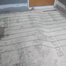 Przygotowanie podłoża pod ogrzewanie podłogowe z rozłożonym zielonym przewodem grzewczym na warstwie kleju z widocznym logo producenta.