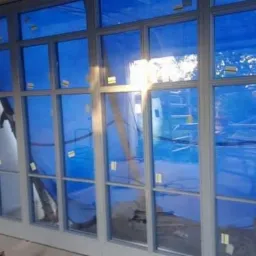 Wnętrze w trakcie prac wykończeniowych; widoczne aluminiowe ramy okienne z niebieską folią ochronną, ściany w stanie surowym, fragment drabiny w tle.