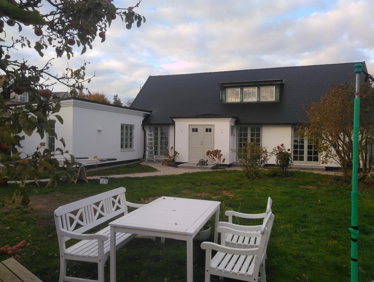 Biały dom z ciemnym dachem widziany z ogrodu, z białymi meblami ogrodowymi na pierwszym planie i drabiną opartą o ścianę budynku.