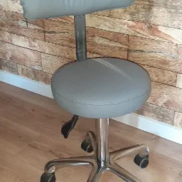 Szary, obrotowy taboret z oparciem na chromowanej podstawie z kółkami, ustawiony na drewnianej podłodze na tle ściany imitującej cegły.