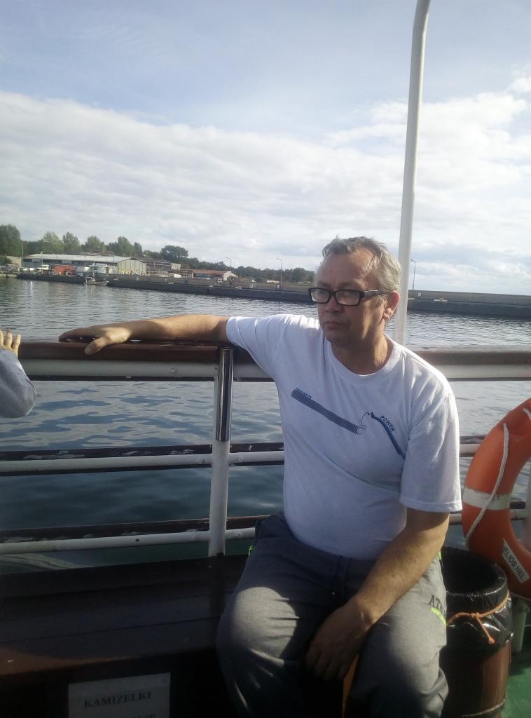 Mężczyzna w okularach siedzi na ławce na promie, w tle woda i budynki portowe.