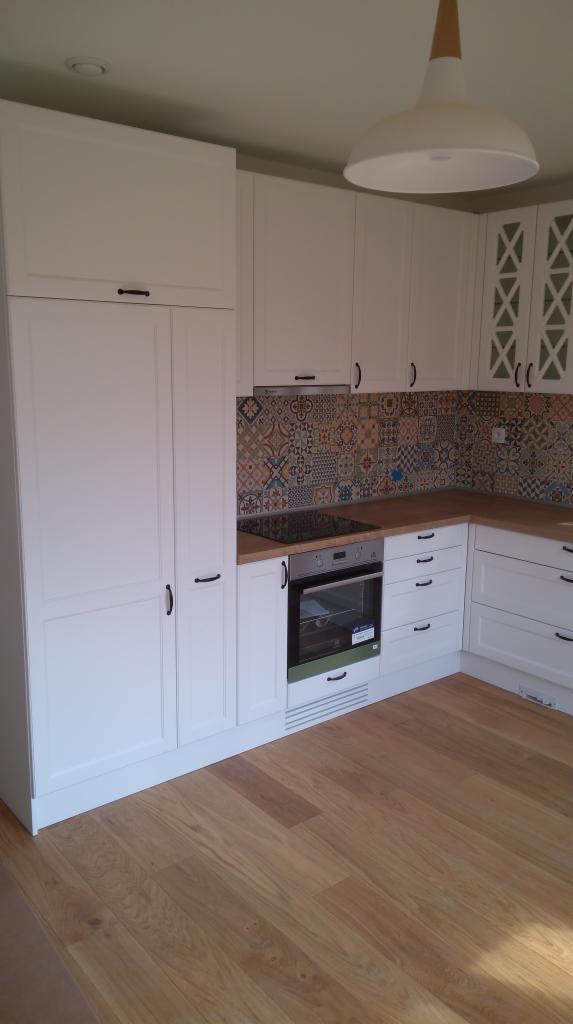 Biała zabudowa kuchenna z drewnianym blatem i kolorowymi płytkami nad blatem, widok na piekarnik i fragment drewnianej podłogi.