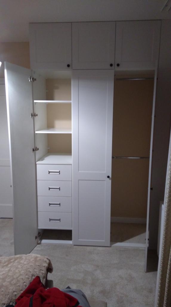 Biała szafa wnękowa z otwartymi drzwiami, widoczne półki, szuflady i drążek na ubrania, wnętrze w kolorze kremowym, na podłodze dywan, w tle ściana w kolorze beżowym.