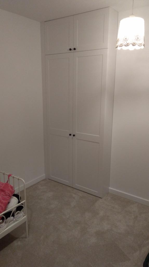 Biała szafa wnękowa z czarnymi gałkami, dopasowana do wysokości pomieszczenia, z widocznym fragmentem łóżeczka dziecięcego i lampą z ażurowym abażurem.