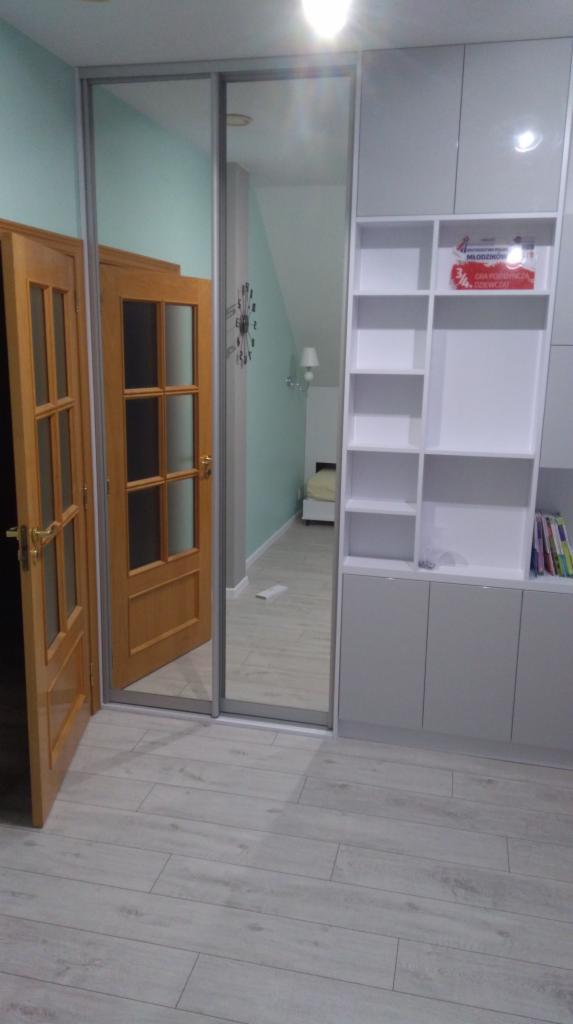 Jasnoszara szafa wnękowa z lustrzanymi drzwiami przesuwnymi, obok drewniane drzwi z szybkami, na podłodze panele w odcieniach szarości.