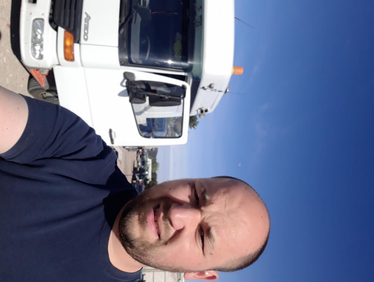 Autoportret mężczyzny na tle białej lawety z pomarańczowym światłem ostrzegawczym, błękitne niebo w tle. Zdjęcie obrócone.