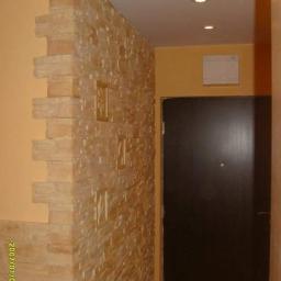 Korzeń & Korzeń Service - Wykończenie korytarza z dekoracyjną ścianą z kamienia w odcieniach beżu, z wkomponowanymi płaskorzeźbami, sąsiadującą z pomalowaną na żółto ścianą i ciemnymi drzwiami.