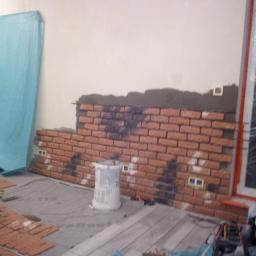 Korzeń & Korzeń Service - Montaż dekoracyjnej ściany z cegły, widoczny etap klejenia cegieł do ściany, wiadro po kleju, częściowo zabezpieczona folią podłoga i ściana.