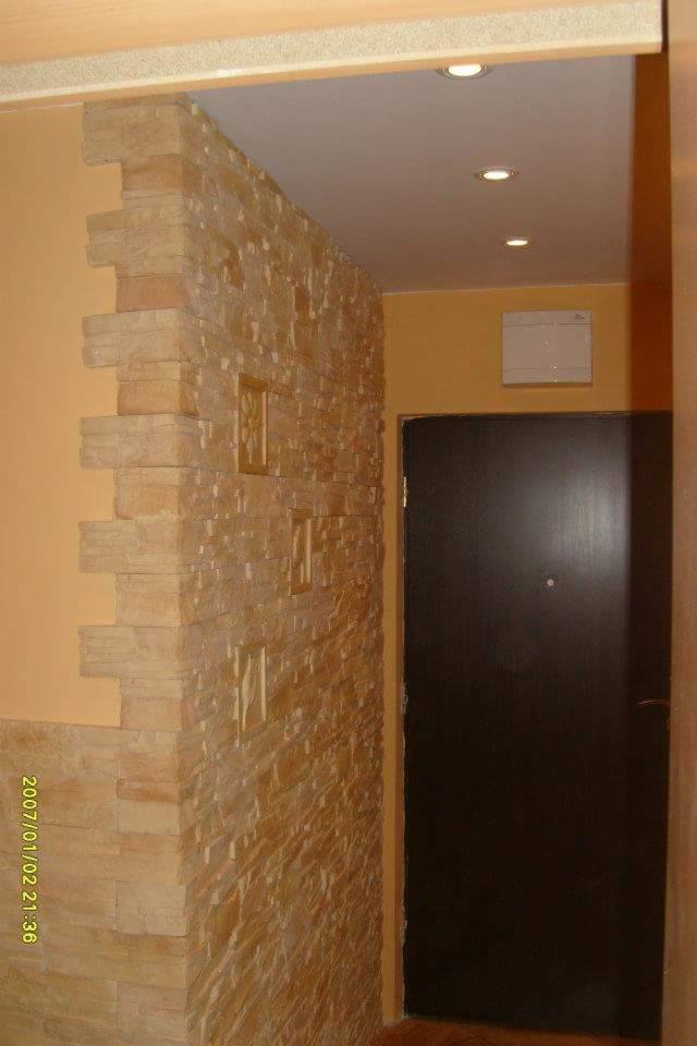 Wykończenie korytarza z dekoracyjną ścianą z kamienia w odcieniach beżu, z wkomponowanymi płaskorzeźbami, sąsiadującą z pomalowaną na żółto ścianą i ciemnymi drzwiami.