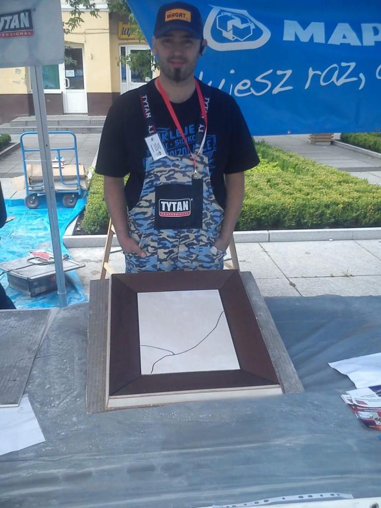 Mężczyzna w kamuflażowym kombinezonie prezentuje ramkę z pękniętą płytką ceramiczną na zewnątrz, na tle baneru reklamowego.