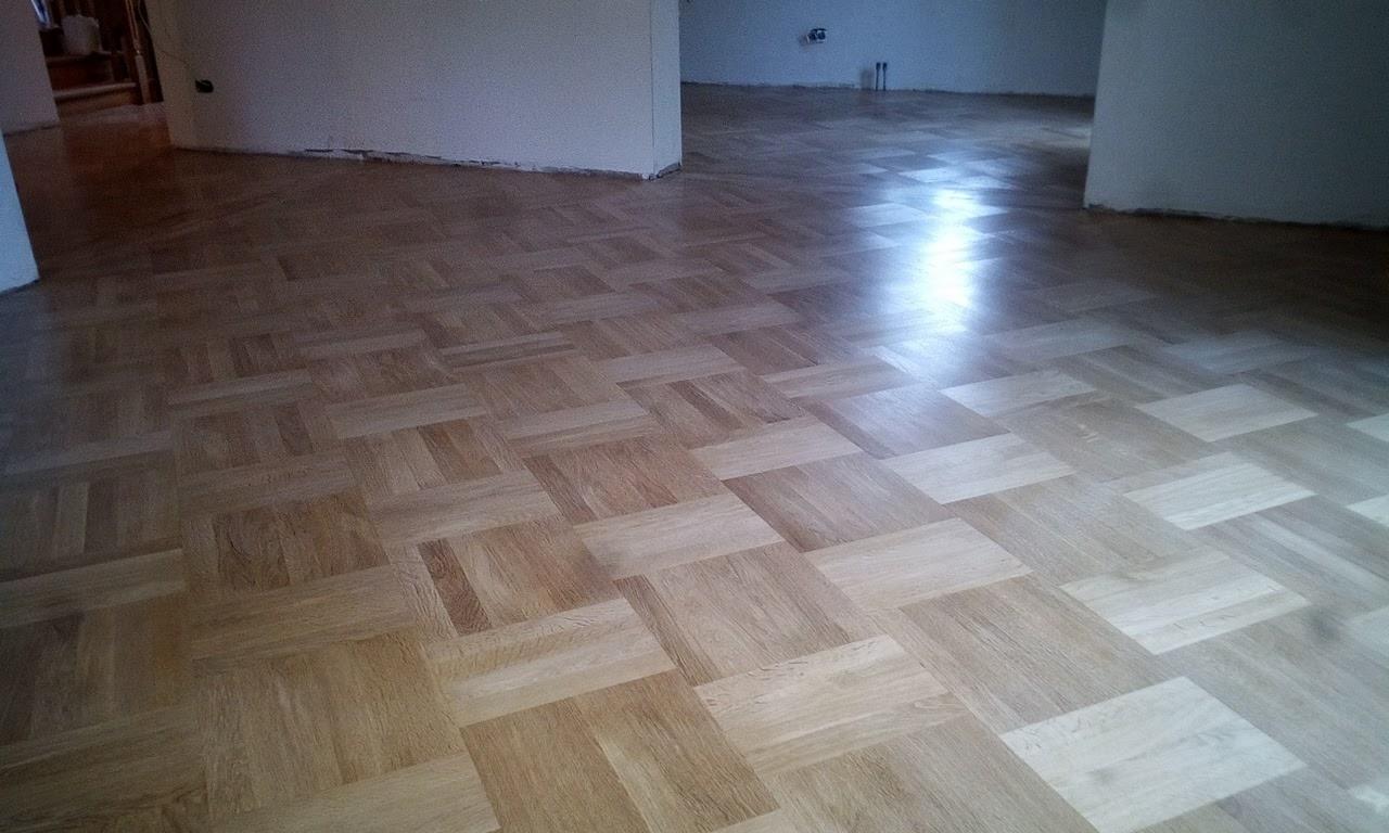Parkiet ułożony we wzór szachownicy, świeżo polakierowany, odbijający światło, widoczne niedoskonałości ściany w tle.