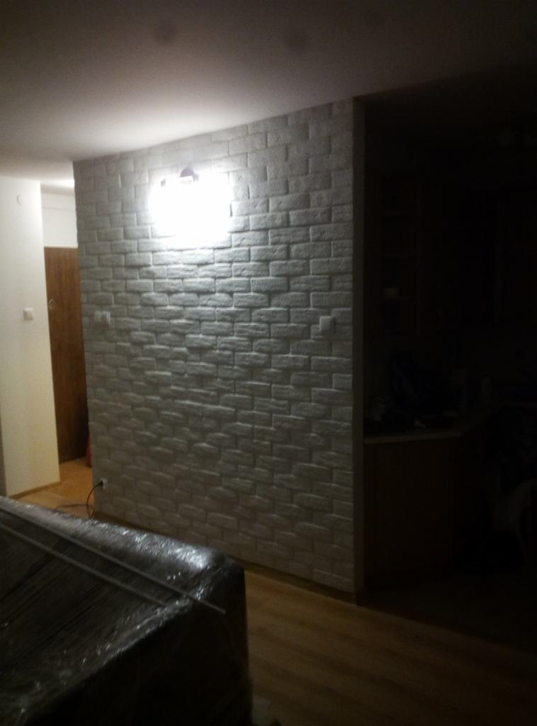 Wykończona ściana w salonie z dekoracyjną cegłą w odcieniach szarości, oświetlona punktowym światłem sufitowym, widoczny fragment podłogi i mebla w folii.