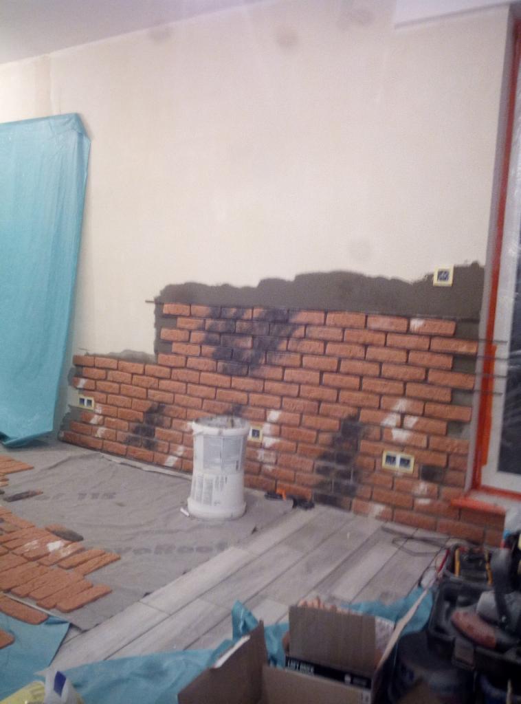 Montaż dekoracyjnej ściany z cegły, widoczny etap klejenia cegieł do ściany, wiadro po kleju, częściowo zabezpieczona folią podłoga i ściana.