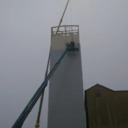 Wznoszenie wysokiej konstrukcji stalowej w Pogorzeli, widoczny montaż obudowy z płyt warstwowych z użyciem podnośnika koszowego i dźwigu. W tle budynek gospodarczy.