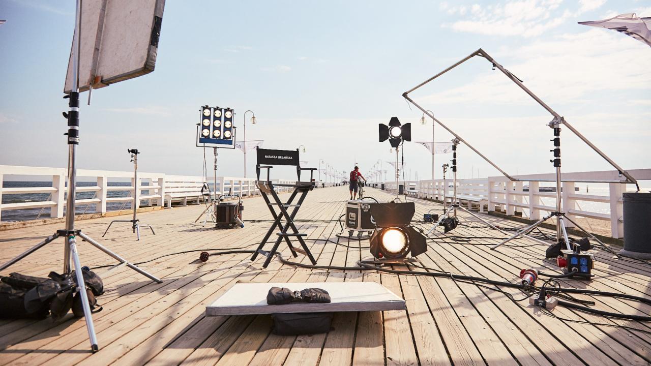 Plan zdjęciowy na molo: reżyserskie krzesło z napisem 'Natalia Urbańska', oświetlenie studyjne, dyfuzory, kable i akcesoria, w tle morze i rowerzysta.