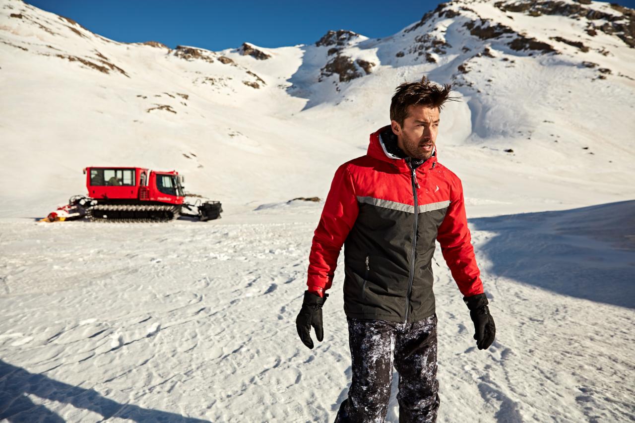 Mężczyzna w czerwono-czarnej kurtce na tle ośnieżonych gór i ratraka w Gdyni, spoglądający w dal.