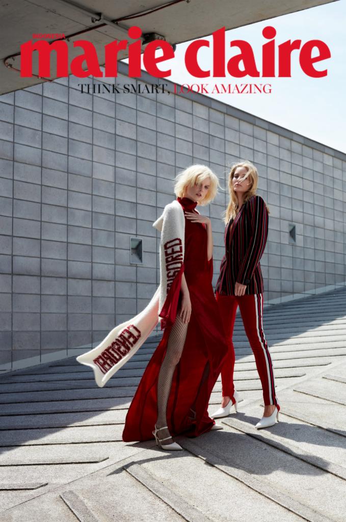 Dwie modelki w czerwonych stylizacjach pozują na tle modernistycznej ściany, jedna w długiej sukni z rozcięciem i szalu z napisem, druga w garniturze w paski z białymi lampasami.