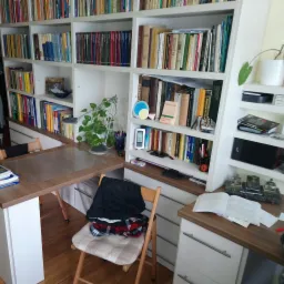 Kącik do pracy z biurkiem i regałem na książki w jasnych kolorach, na krześle leżą ubrania.