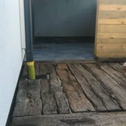 Drewniana podłoga z belek w Radomiu, z odpływem liniowym przy ścianie budynku z szorstkim tynkiem, obok ściany z desek i sterta drewna.