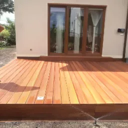 Nowoczesny drewniany taras z wbudowanym oświetleniem, przylegający do budynku z brązowymi oknami; w tle podlewanie trawnika.