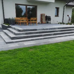 Nowoczesny taras z szarej kostki brukowej, schody prowadzące do domu, drewniane meble ogrodowe, zadbany trawnik, element zagospodarowania terenu wokół domu.