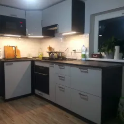 Nowoczesna kuchnia z białymi szafkami i czarnymi akcentami, widok na okno z roślinami doniczkowymi, oświetlenie podszafkowe.