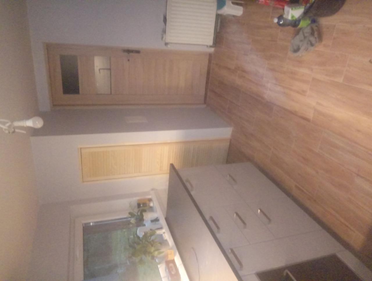 Jasna łazienka z drewnianymi akcentami: płytki imitujące drewno na podłodze i ścianie, drewniane drzwi z szybą, biała szafka pod umywalkę, okno z roślinami. Widoczne niedoskonałości i brak porządku.