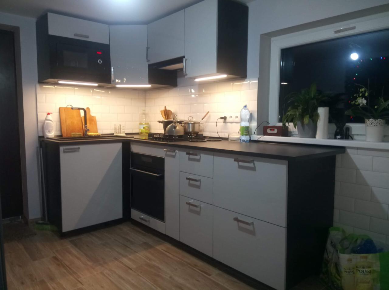 Nowoczesna kuchnia z białymi szafkami i czarnymi akcentami, widok na okno z roślinami doniczkowymi, oświetlenie podszafkowe.