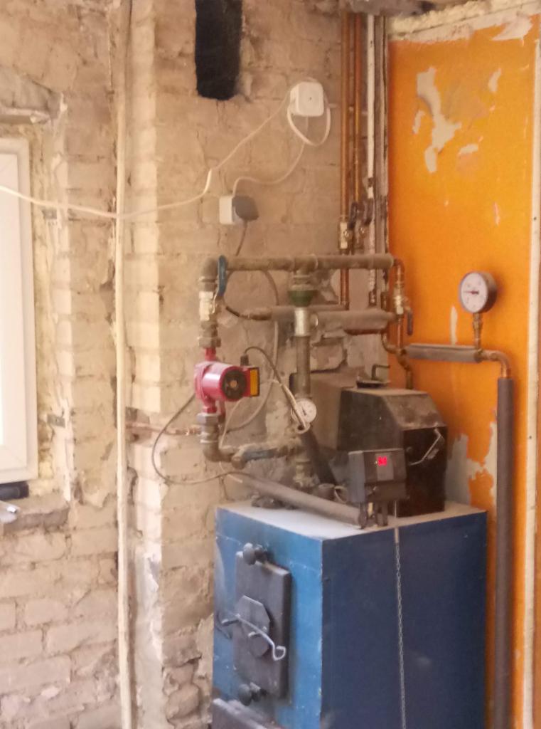 Stara kotłownia z piecem na paliwo stałe, widoczne rury, zawory, manometry, ściana z cegły i fragment pomarańczowych drzwi z resztkami farby.