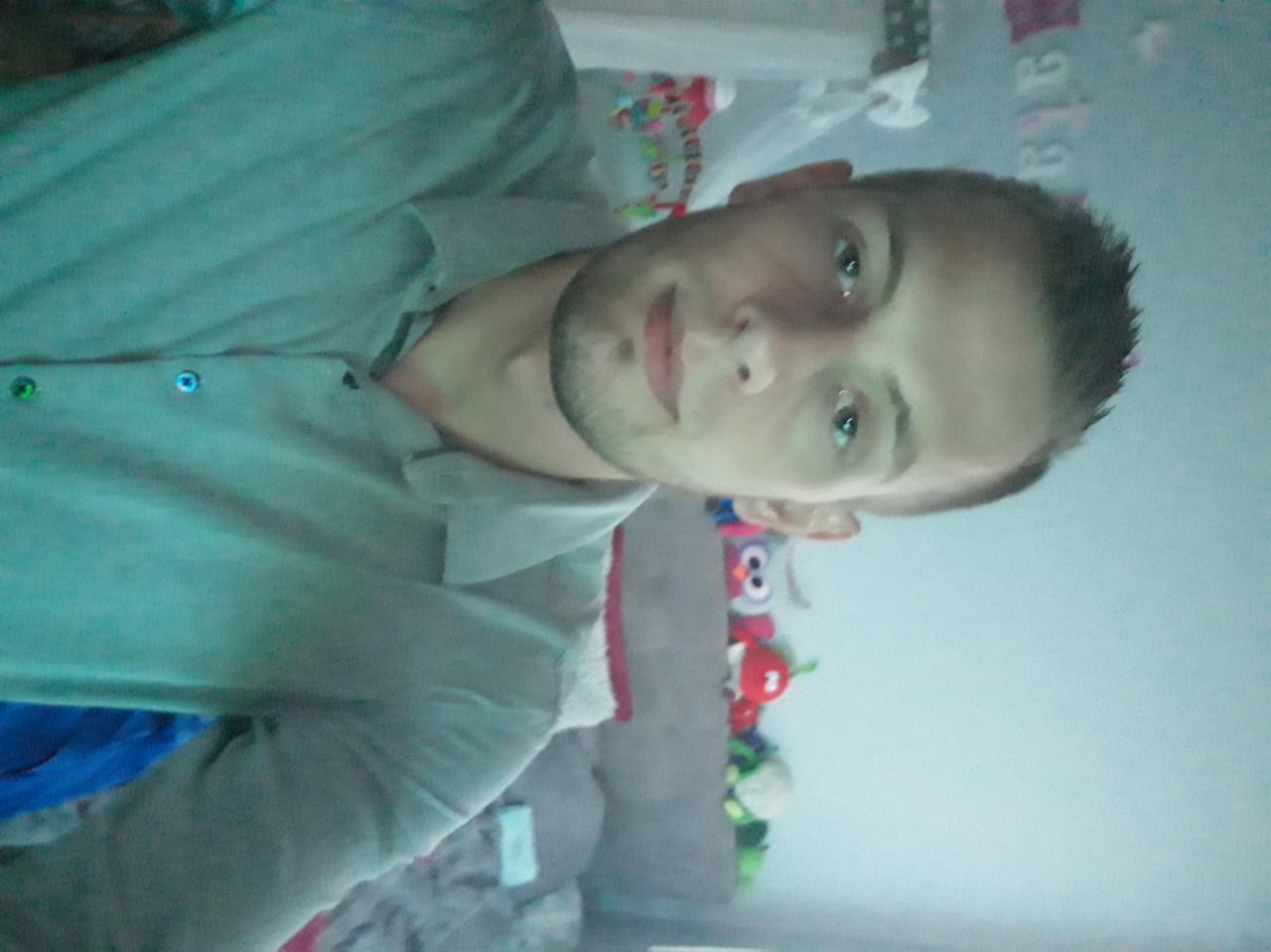 Młody mężczyzna w szarej koszuli, z lekkim zarostem, patrzy w obiektyw. W tle widać ścianę i pluszowe zabawki. Zdjęcie obrócone, nieostre.