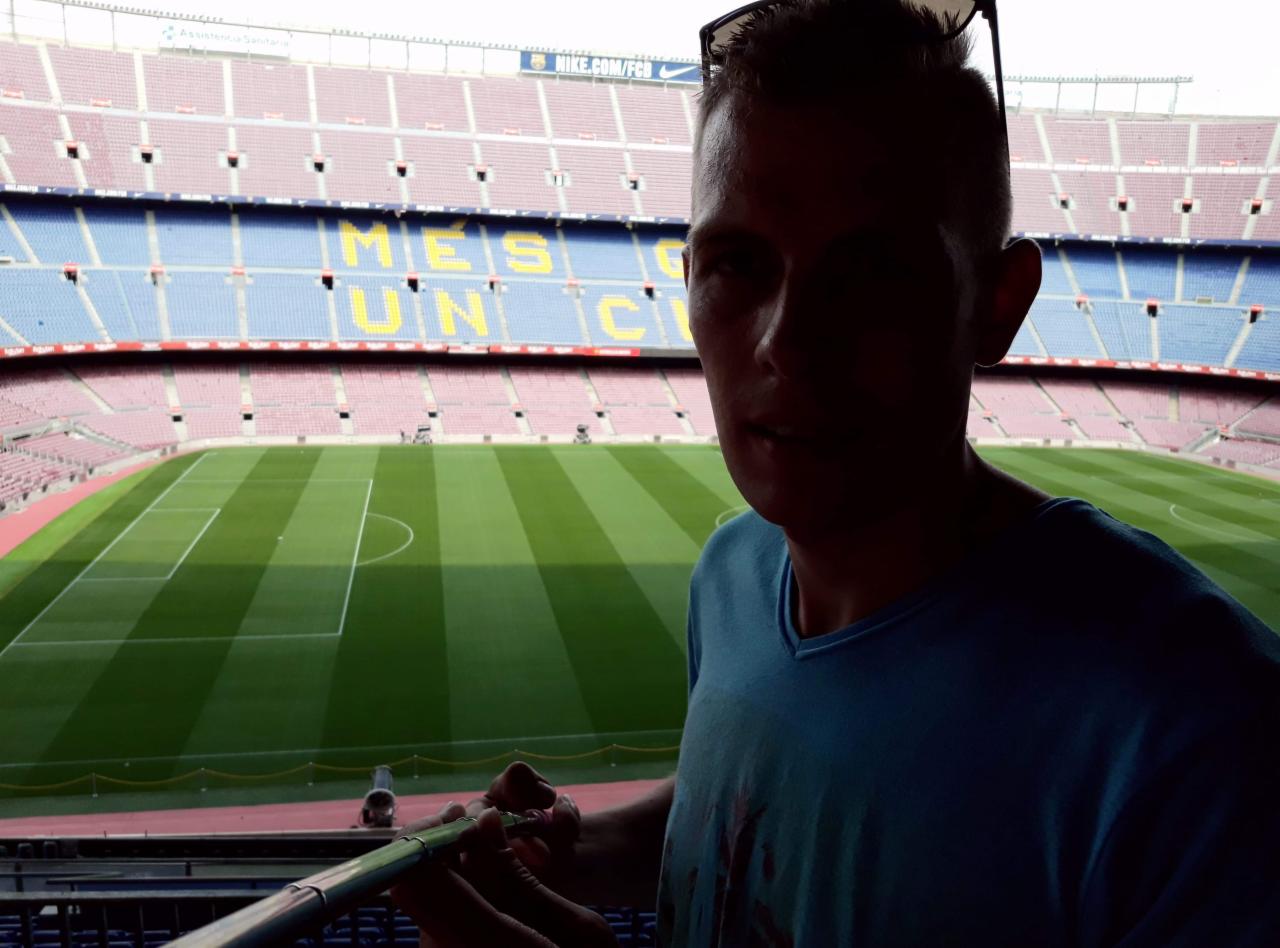 Mężczyzna na tle stadionu Camp Nou w Barcelonie, częściowo zacieniony, z widocznym boiskiem i trybunami. Osoba w koszulce, trzyma coś w rękach.