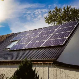 Panele słoneczne zamontowane na dachu domu jednorodzinnego z oknem dachowym, widoczne drzewa i błękitne niebo w tle.