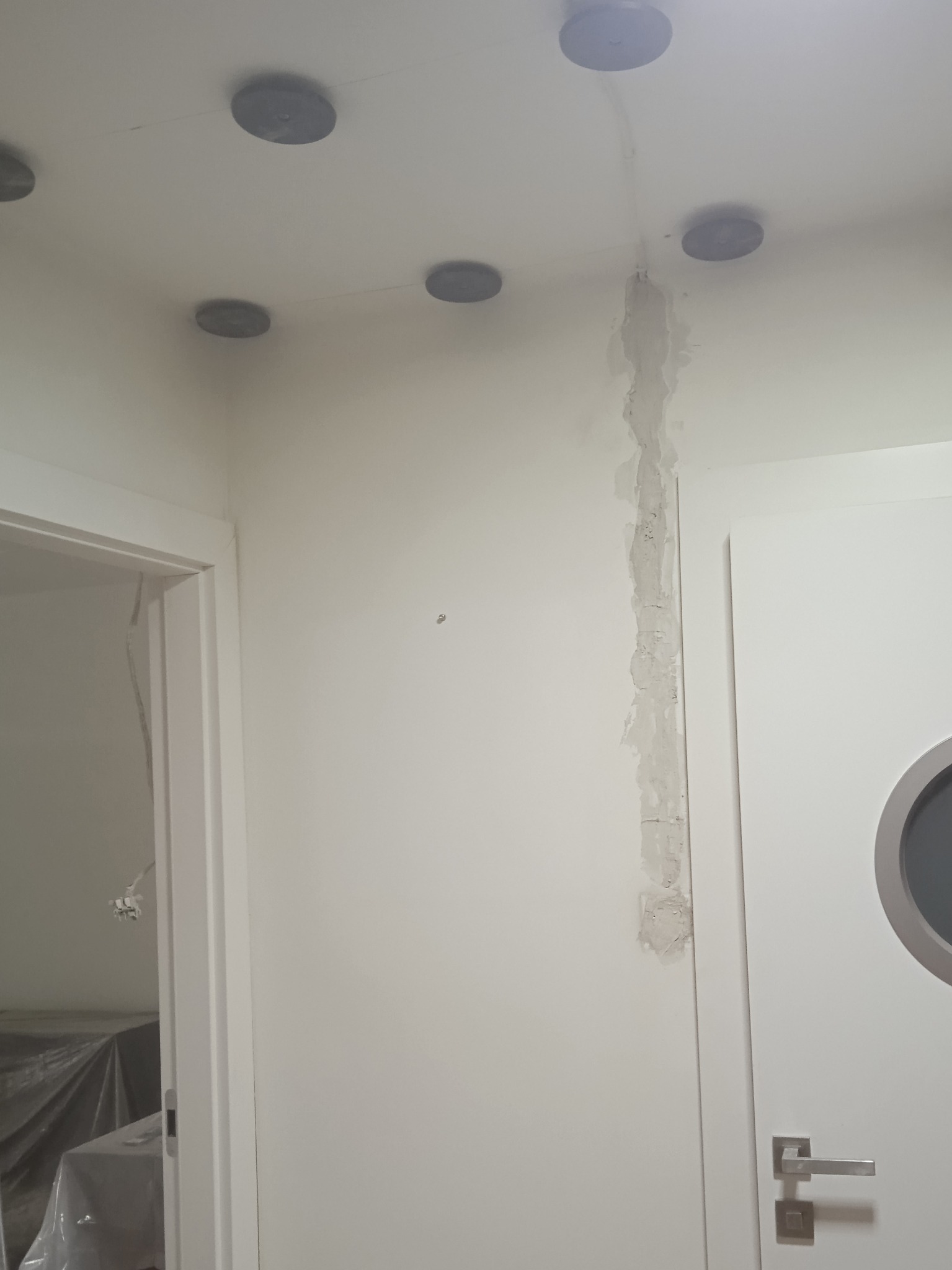 Wnętrze z zainstalowanymi okrągłymi lampami sufitowymi, widoczne ślady po instalacji elektrycznej wymagające wykończenia, białe ściany i drzwi z okrągłym oknem.