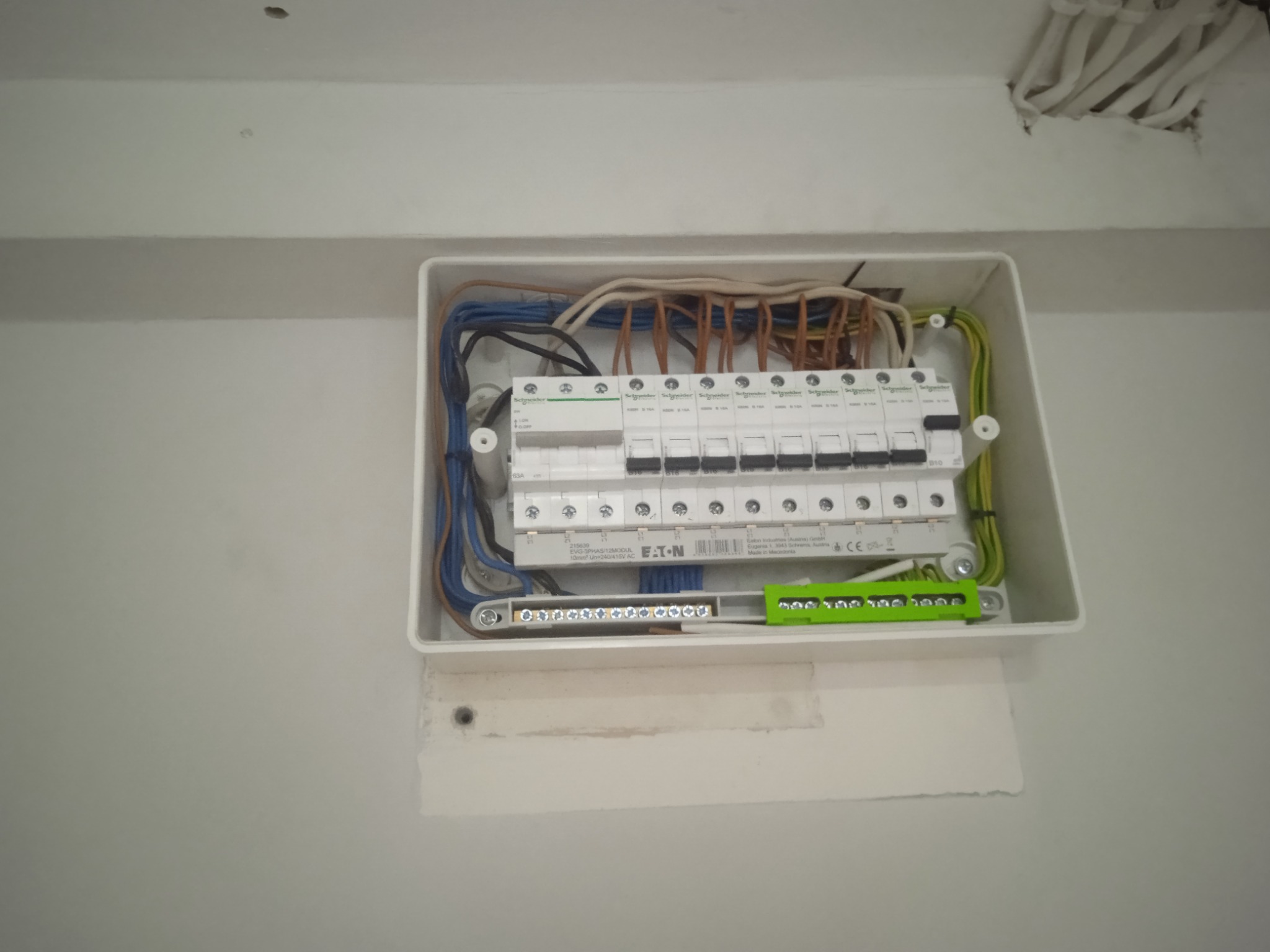 Otwarta skrzynka elektryczna z zainstalowanymi bezpiecznikami Schneider i Eaton, widoczne kable w różnych kolorach, mocowana na białej ścianie wewnątrz budynku.