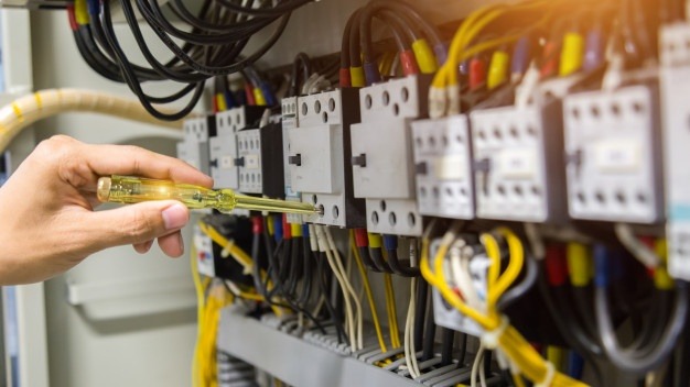 Elektryk sprawdza napięcie w rozdzielnicy za pomocą próbnika neonowego z przezroczystą, żółtą rączką, na tle widoczne bezpieczniki i okablowanie.