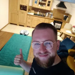 Mężczyzna w okularach i czarnej koszulce robi selfie na tle zielonego stołu do masażu i drewnianej kuchni, pokazując kciuk w górę.