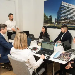 Zesp&oacute;ł w biurze nieruchomości omawia projekt Baltic Wave, widoczny na dużym plakacie za nimi, przy użyciu laptop&oacute;w i notatnik&oacute;w.