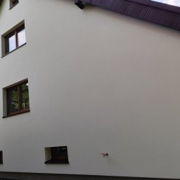 PERSPEKTYWA KOSTIANTYN SOBKO - Jasna elewacja domu jednorodzinnego z brązowymi ramami okiennymi i ciemnobrązowym dachem, widoczne trzy okna na różnych wysokościach i kran na ścianie.