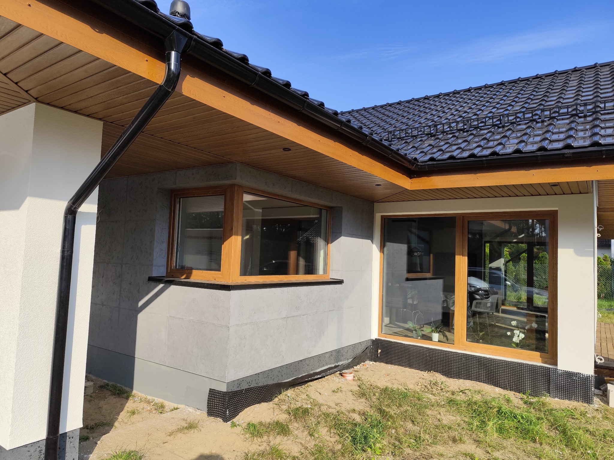 Dom jednorodzinny z szarą elewacją imitującą beton architektoniczny, drewnianą stolarką okienną i czarną rynną, widoczna folia fundamentowa.