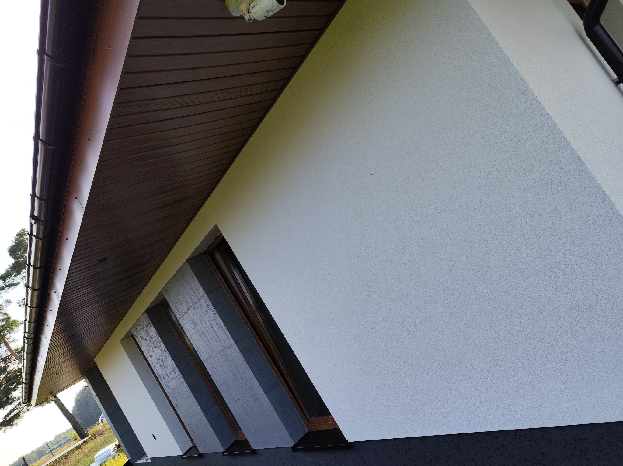 Nowoczesna elewacja domu z tynkiem strukturalnym w jasnym odcieniu, beton architektoniczny na dolnej części ściany, okna z szarymi panelami i brązowymi ramami, oraz ciemnobrązowe podbitki dachowe...